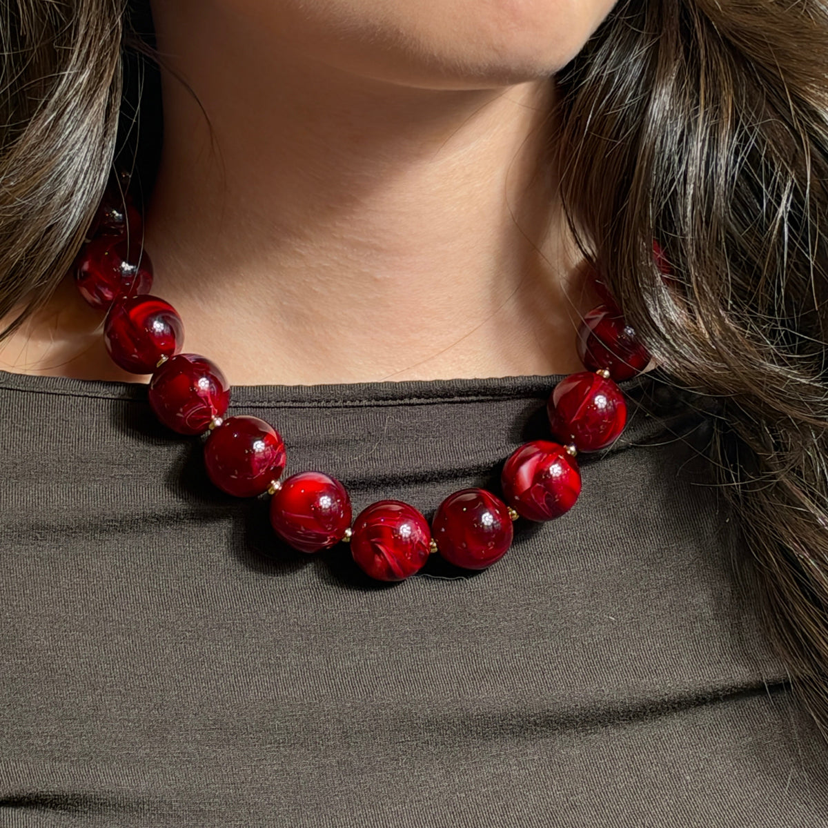 Chunky Statement-Kette mit großen, glänzenden, bordeauxroten Acrylperlen in marmorierter Optik und kleinen goldfarbenen Edelstahlperlen. Die Kette wird am Hals getragen und zu einem dunkelgrauen Oberteil kombiniert.